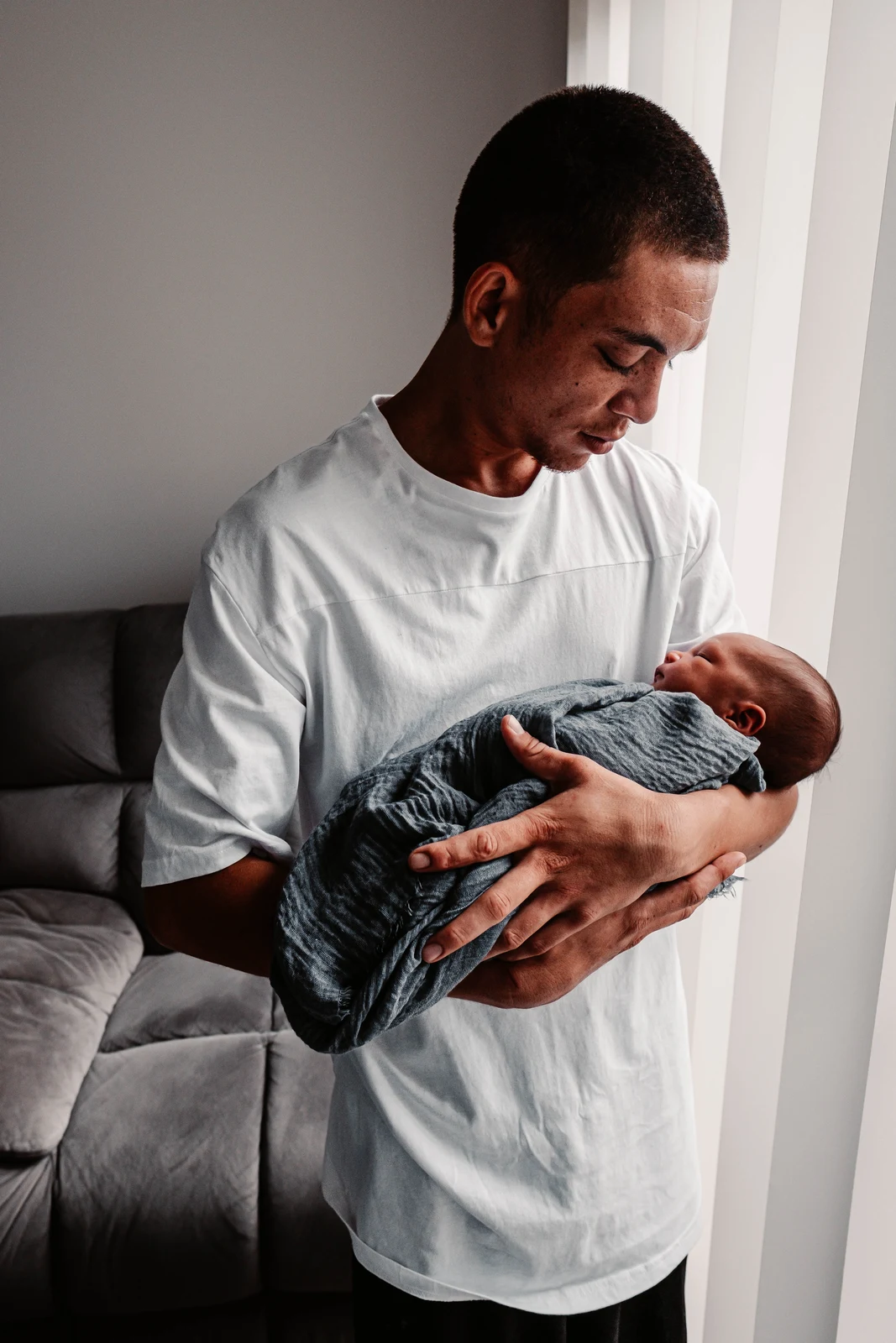 Dad holding his sleeping baby wrapped in a blue-grey blanket beside a window
